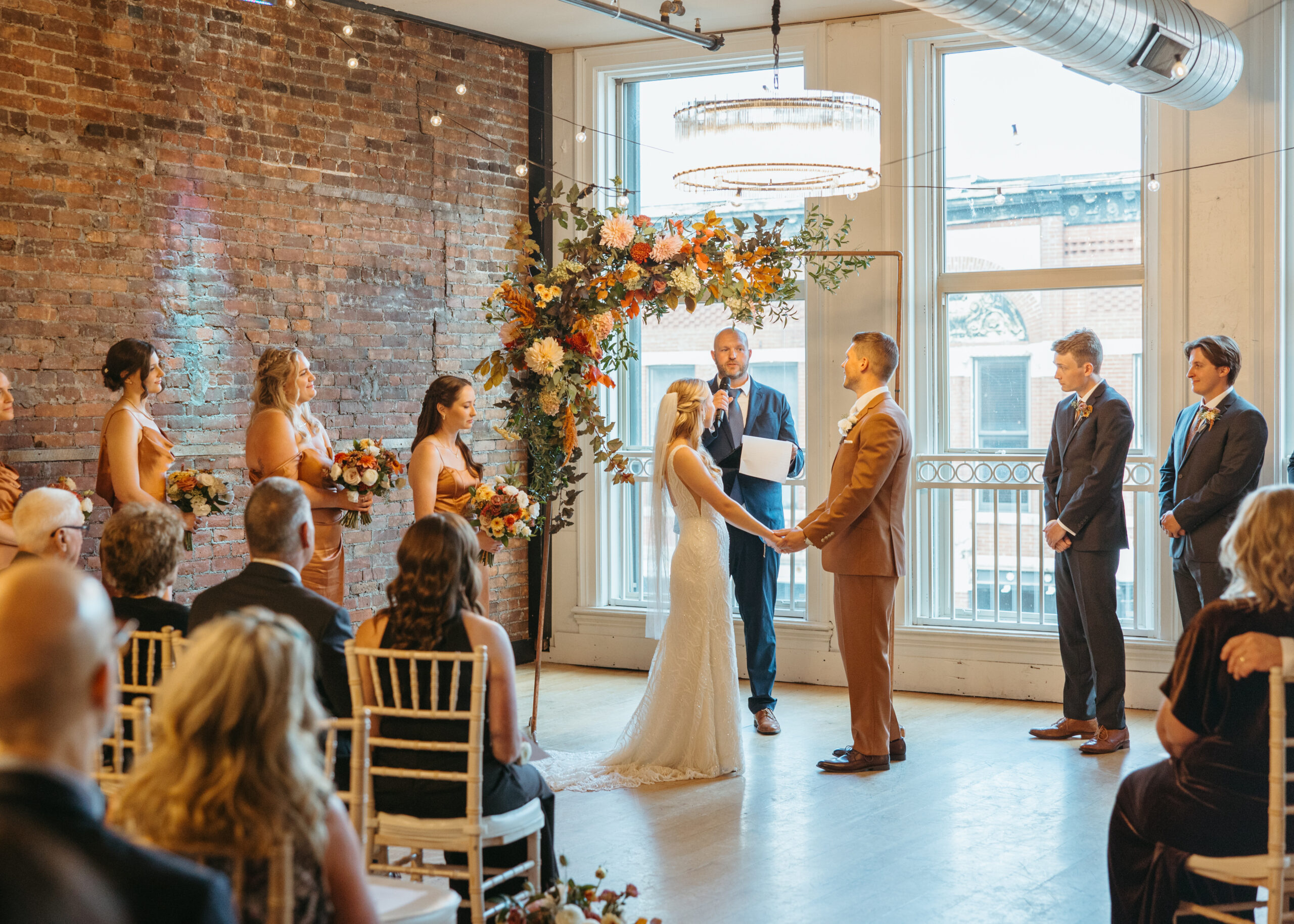A wedding ceremony at Loft 214 in Stillwater, MN, showing a couple standing before a floral arch in front of large industrial windows with views of the St. Croix River.