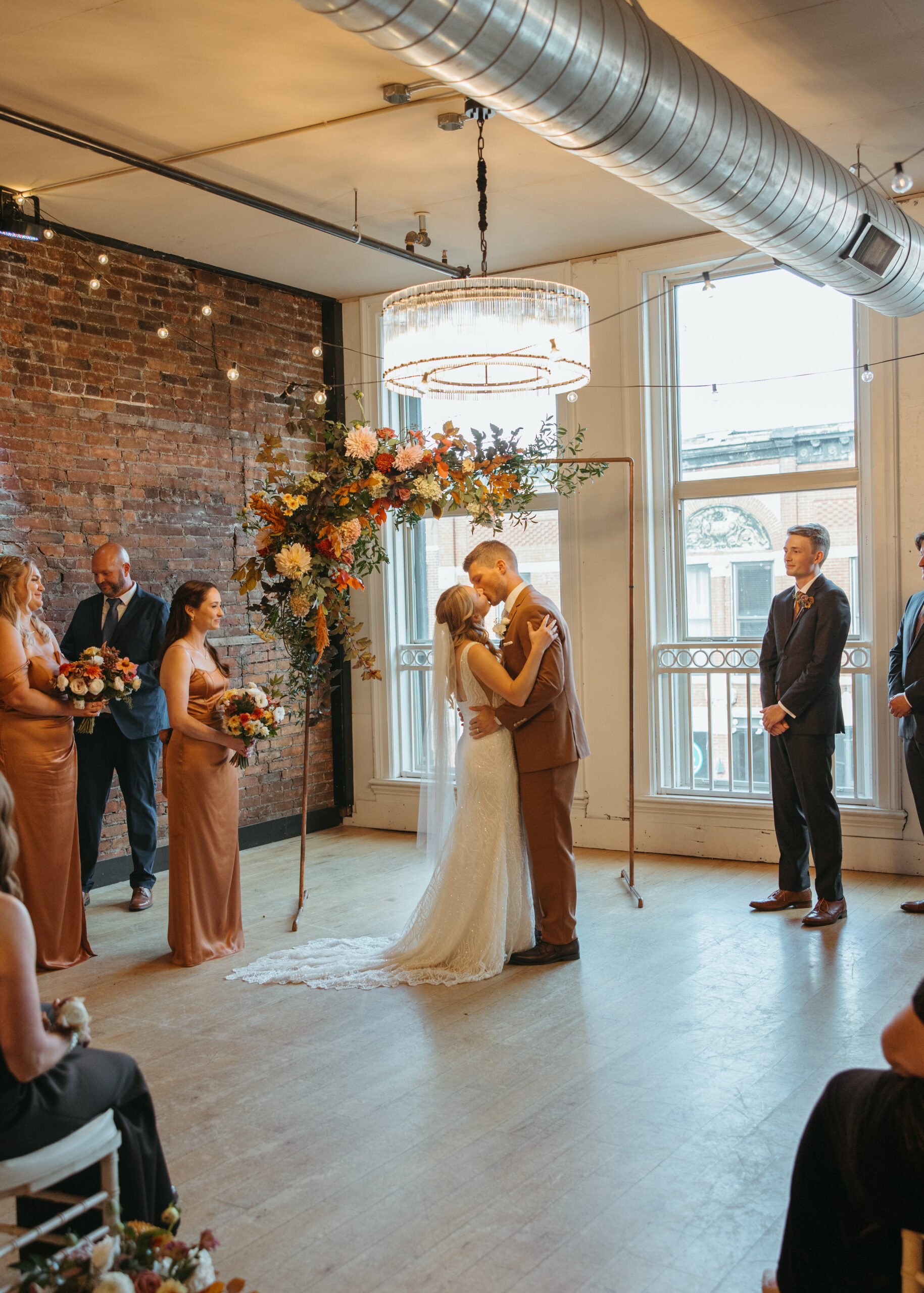 A wedding ceremony at Loft 214 in Stillwater, MN, showing a couple standing before a floral arch in front of large industrial windows with views of the St. Croix River.