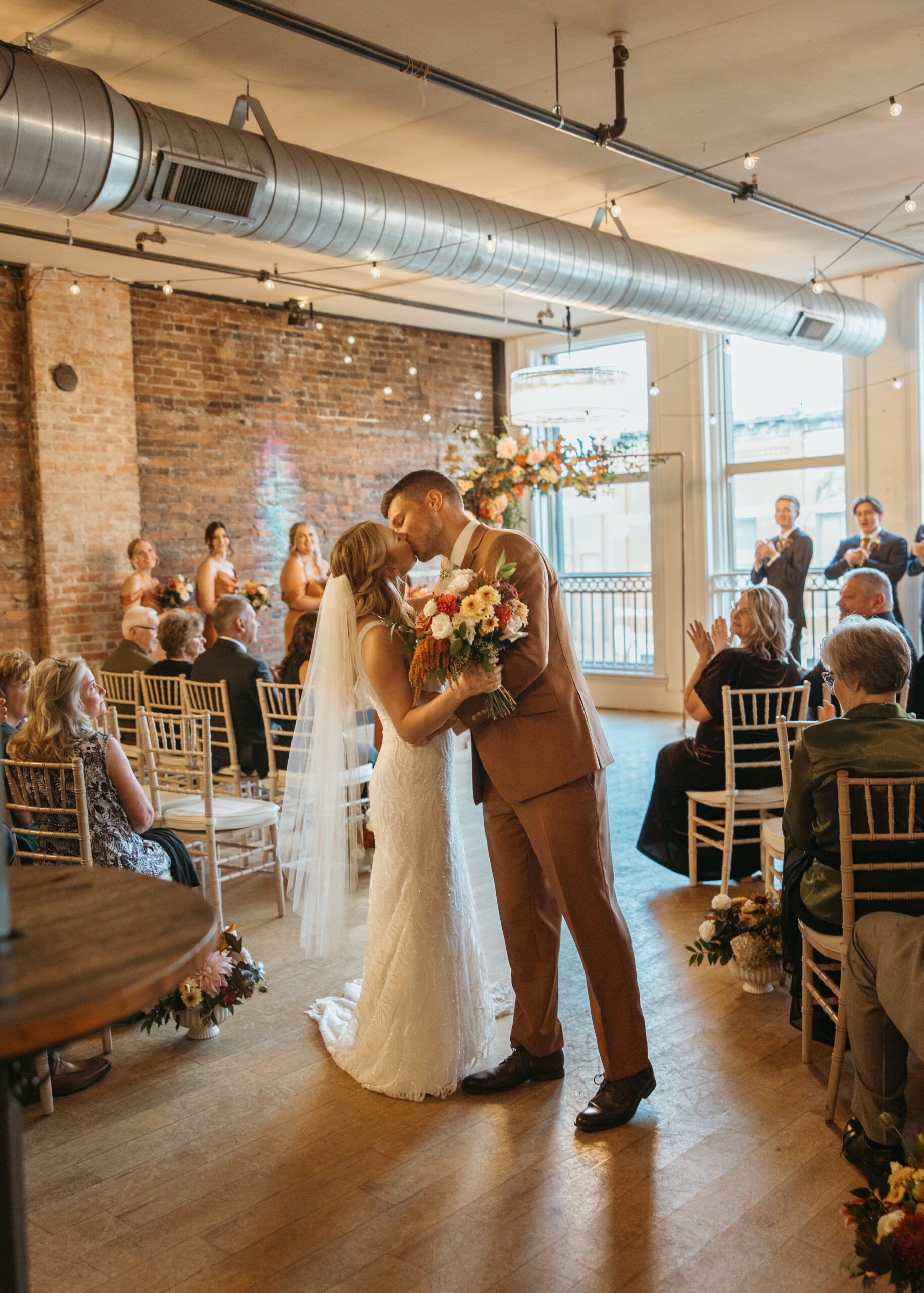 A wedding ceremony at Loft 214 in Stillwater, MN, showing a couple standing before a floral arch in front of large industrial windows with views of the St. Croix River.