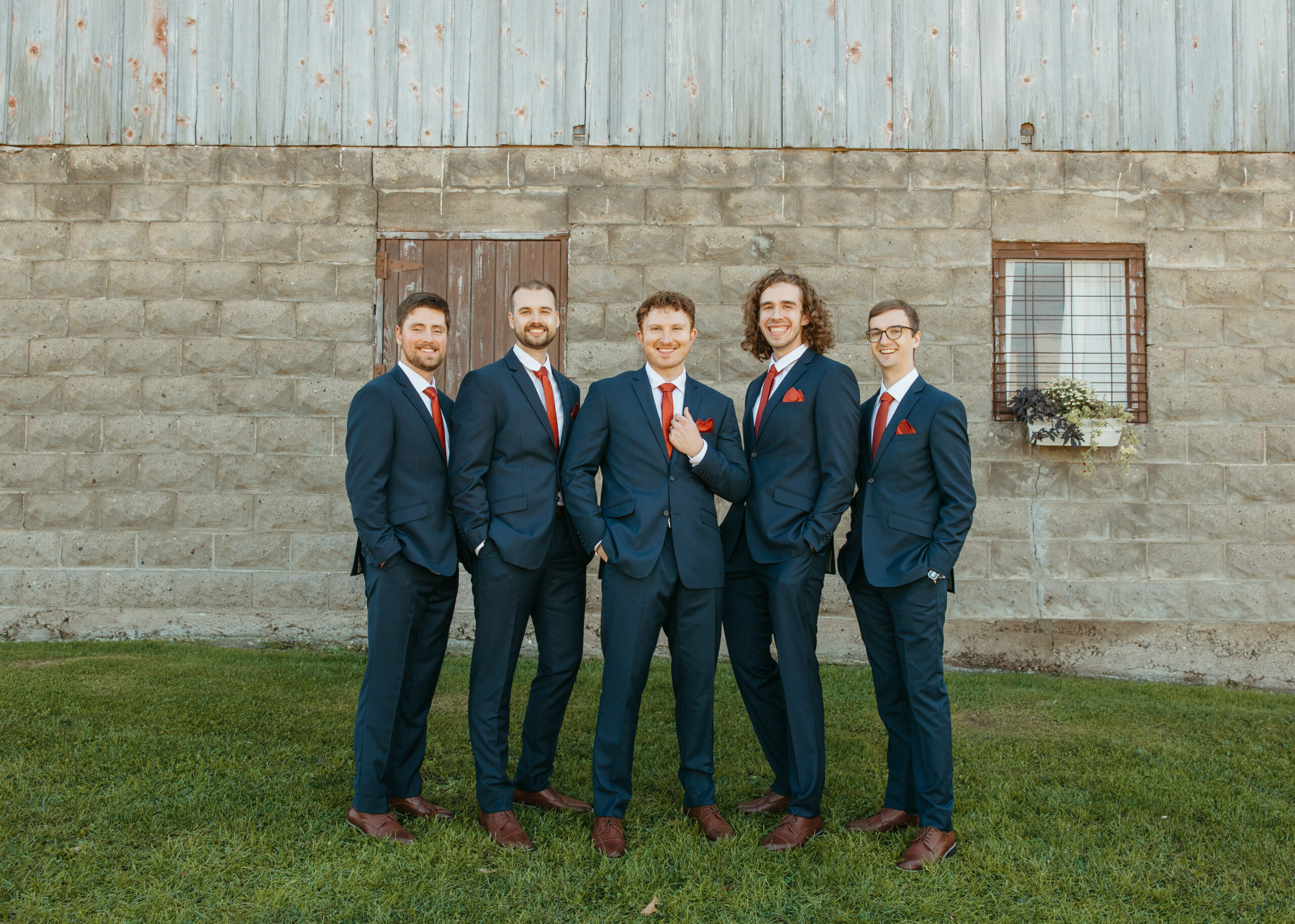 Fun and candid groomsmen portrait with the groom laughing as his friends tease him outside a rustic stone building for a Minnesota wedding.
