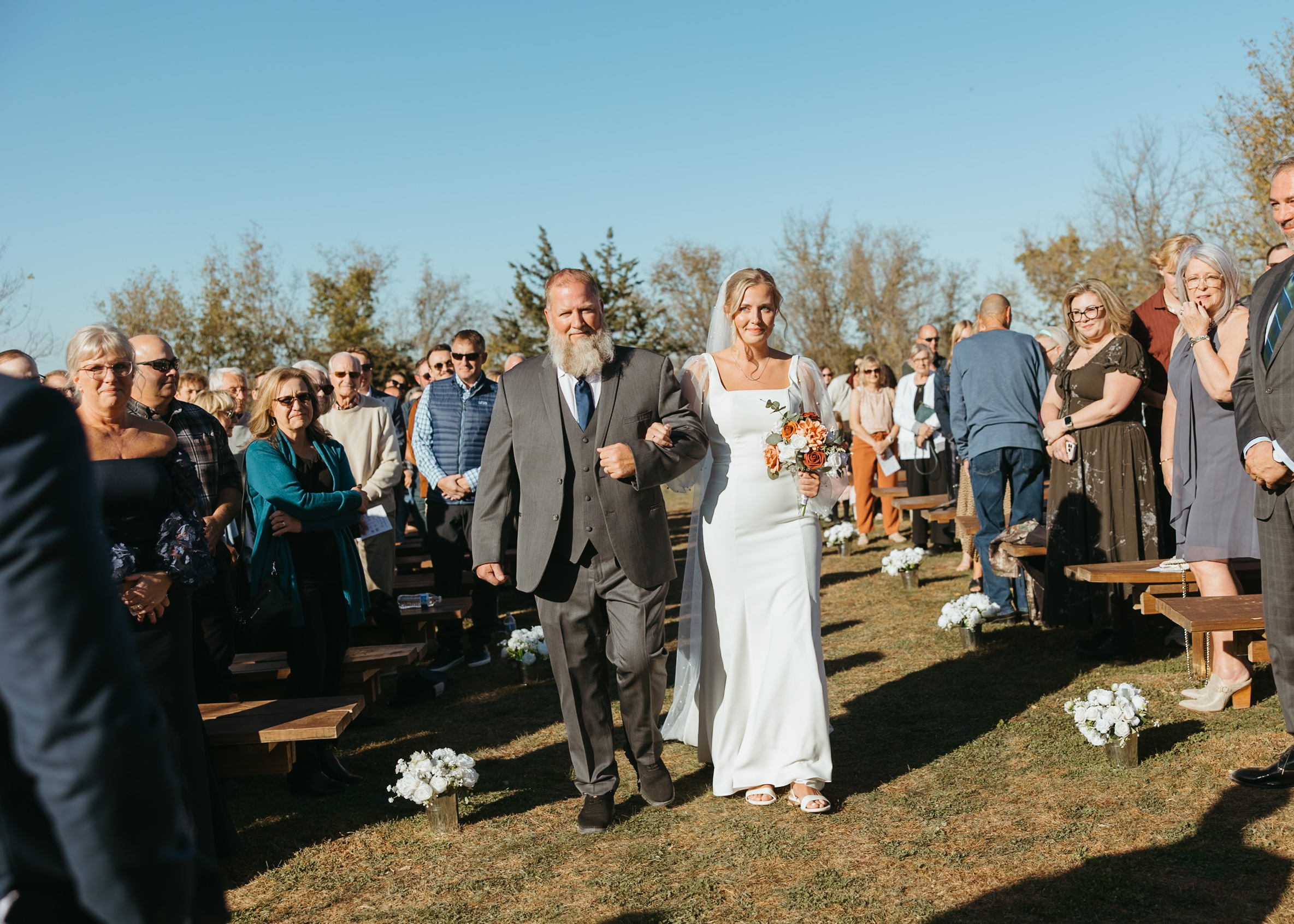 A bride in a fitted long-sleeve pearl wedding dress walking down the aisle with her father at an outdoor Genesis Ranch ceremony, captured by a Minneapolis wedding photographer.