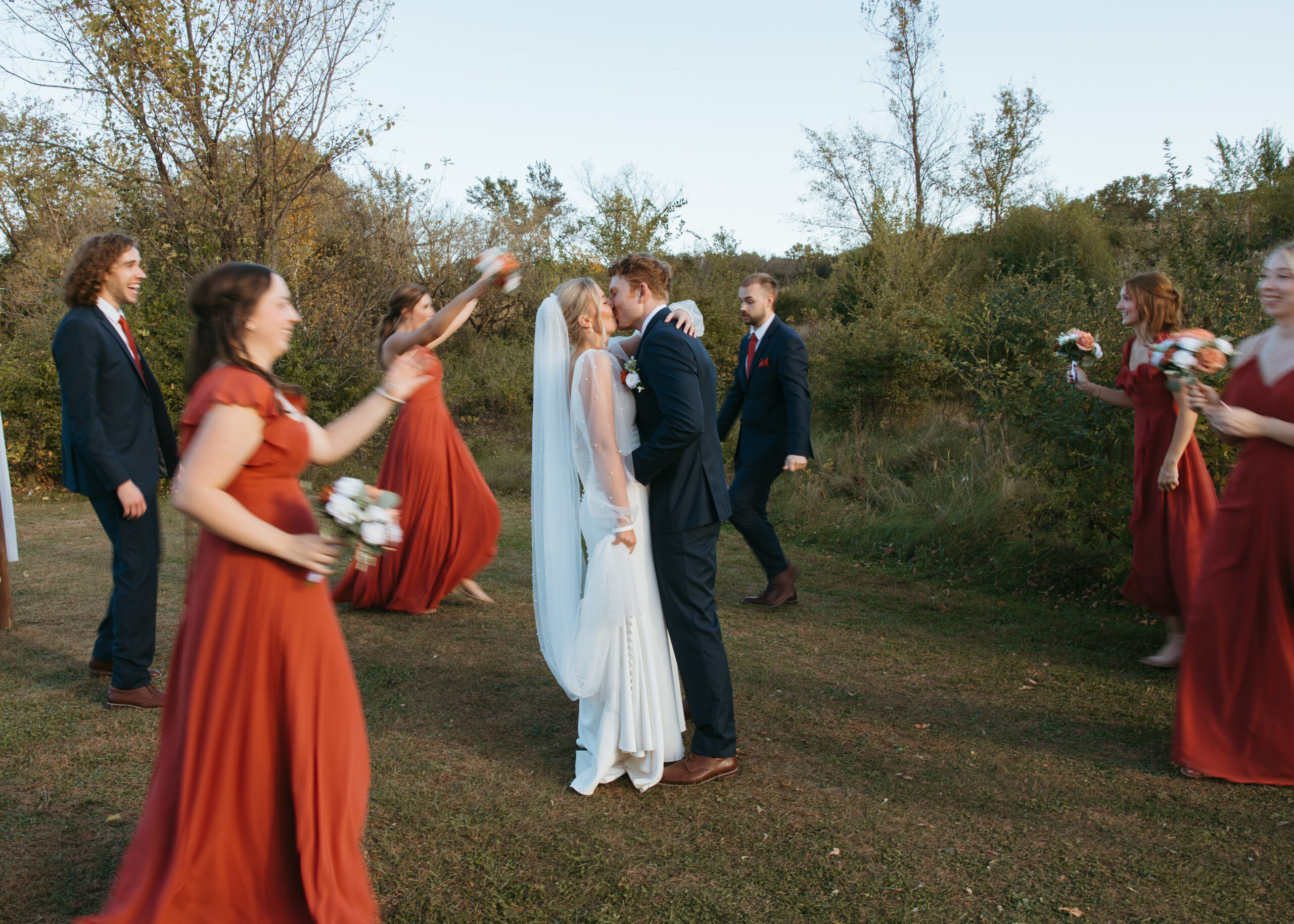 Bride and groom kissing in a field at Genesis Ranch while their wedding party in navy suits and terracotta dresses celebrates around them, captured by a Minneapolis wedding photographer.