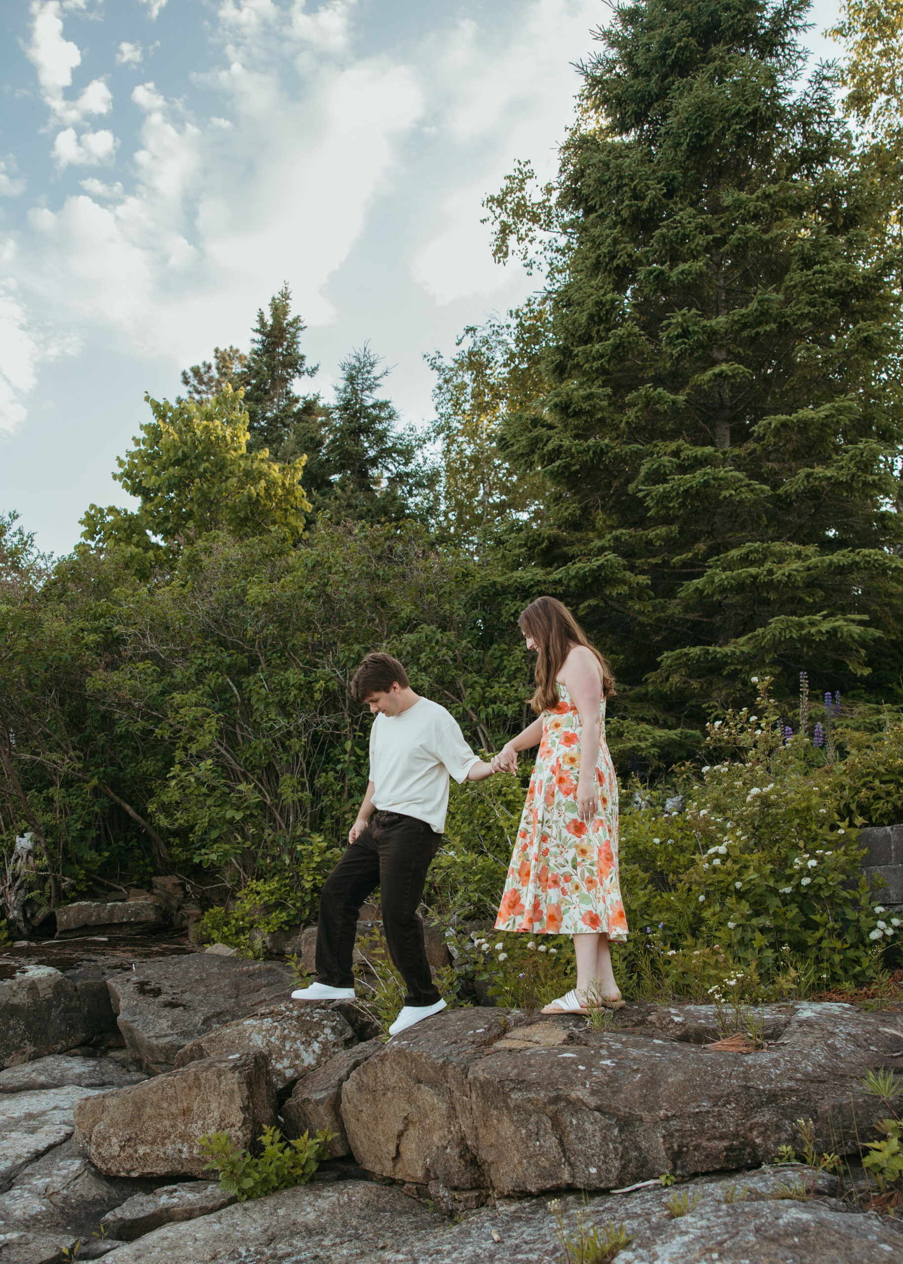 A happy couple standing on the rugged, rocky shoreline of Lake Superior in Lutsen, Minnesota, with waves crashing against the rocks under a bright summer sky.