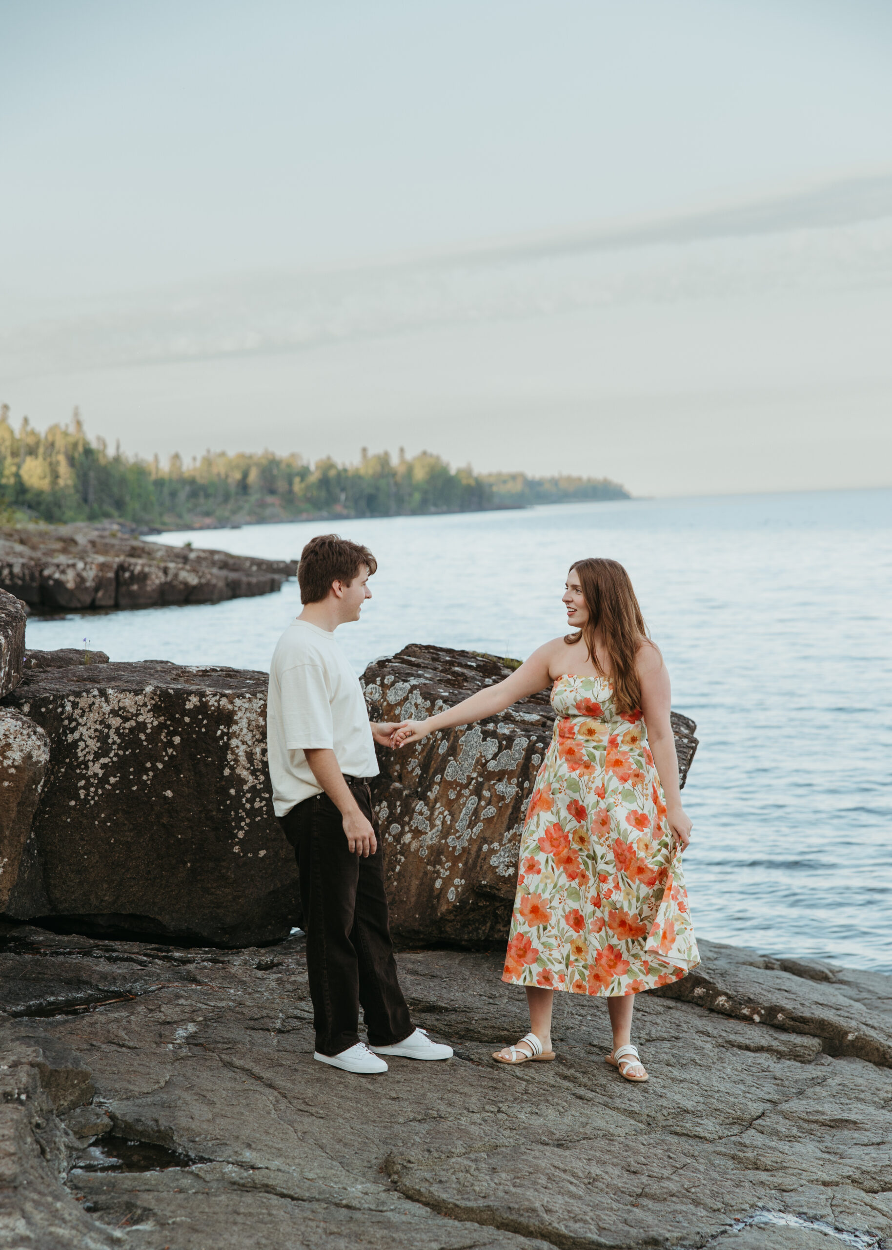 A happy couple standing on the rugged, rocky shoreline of Lake Superior in Lutsen, Minnesota, with waves crashing against the rocks under a bright summer sky.