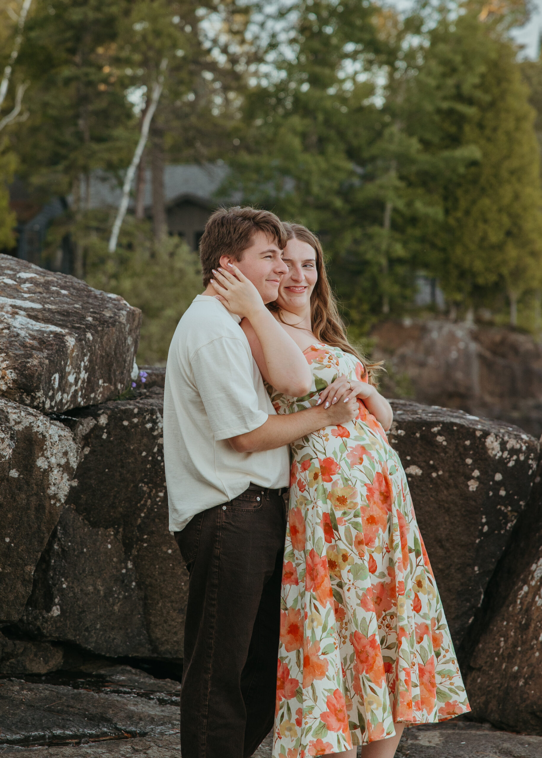 A happy couple standing on the rugged, rocky shoreline of Lake Superior in Lutsen, Minnesota, with waves crashing against the rocks under a bright summer sky.