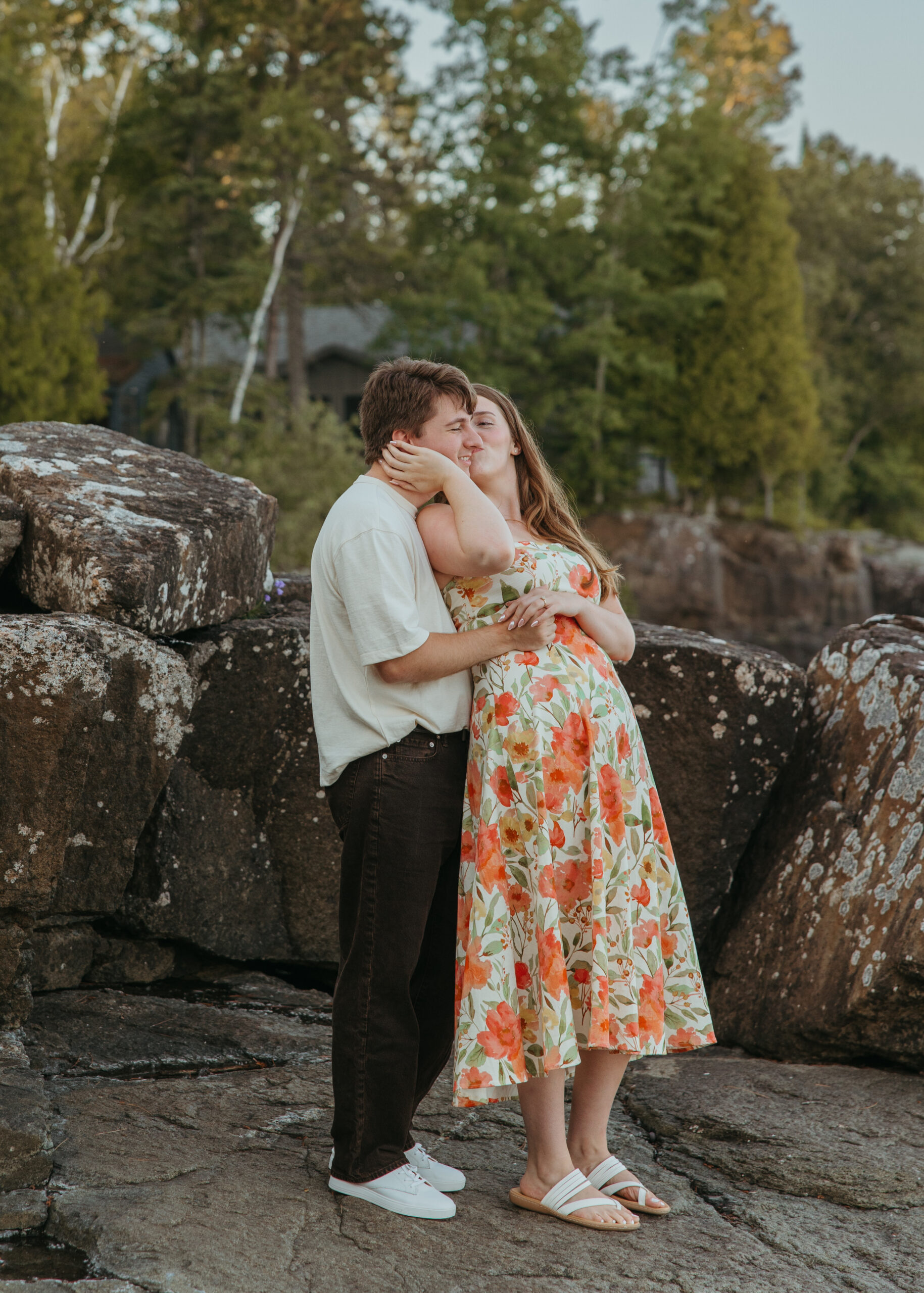 A happy couple standing on the rugged, rocky shoreline of Lake Superior in Lutsen, Minnesota, with waves crashing against the rocks under a bright summer sky.