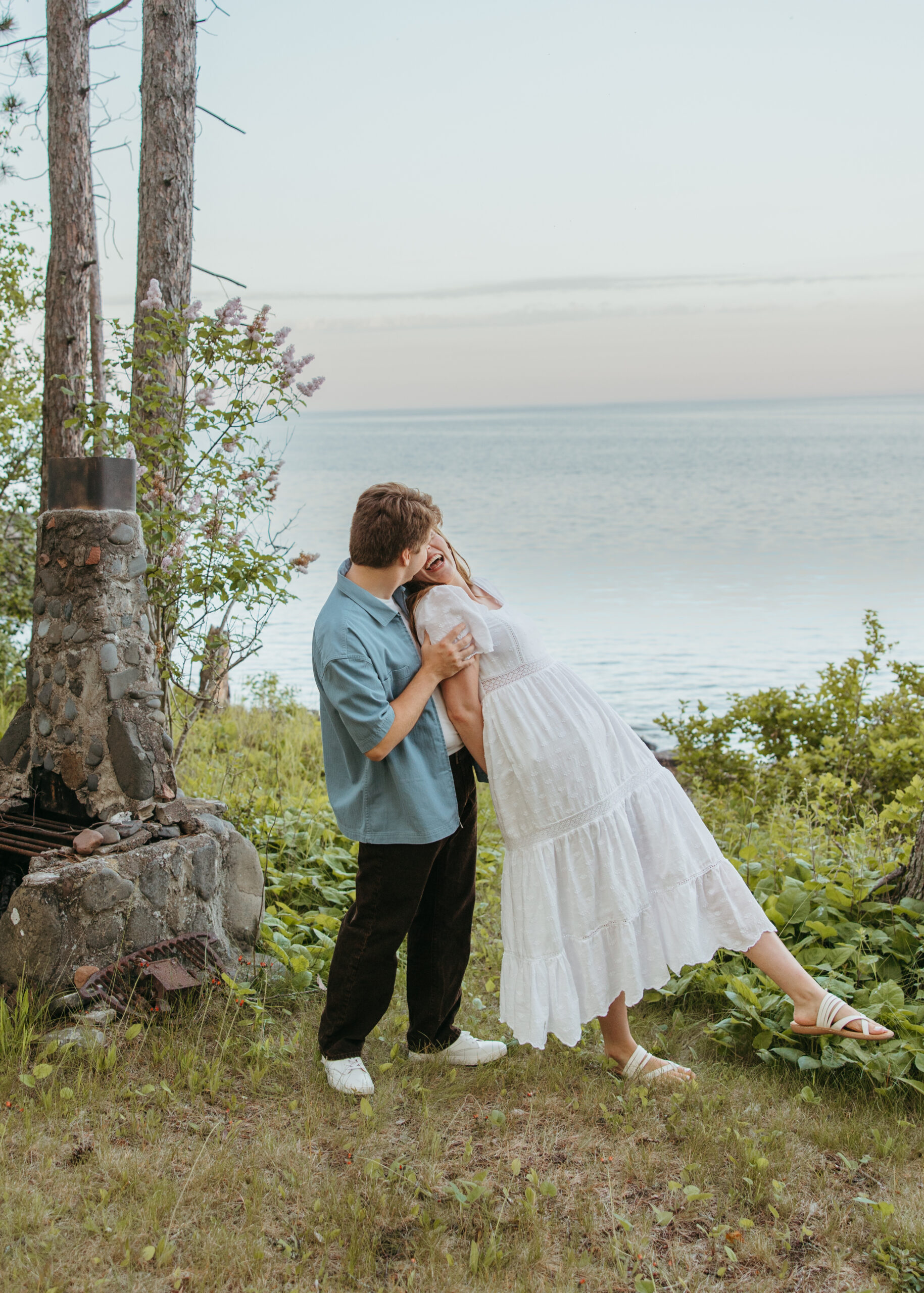 A happy couple standing on the rugged, rocky shoreline of Lake Superior in Lutsen, Minnesota, with waves crashing against the rocks under a bright summer sky.
