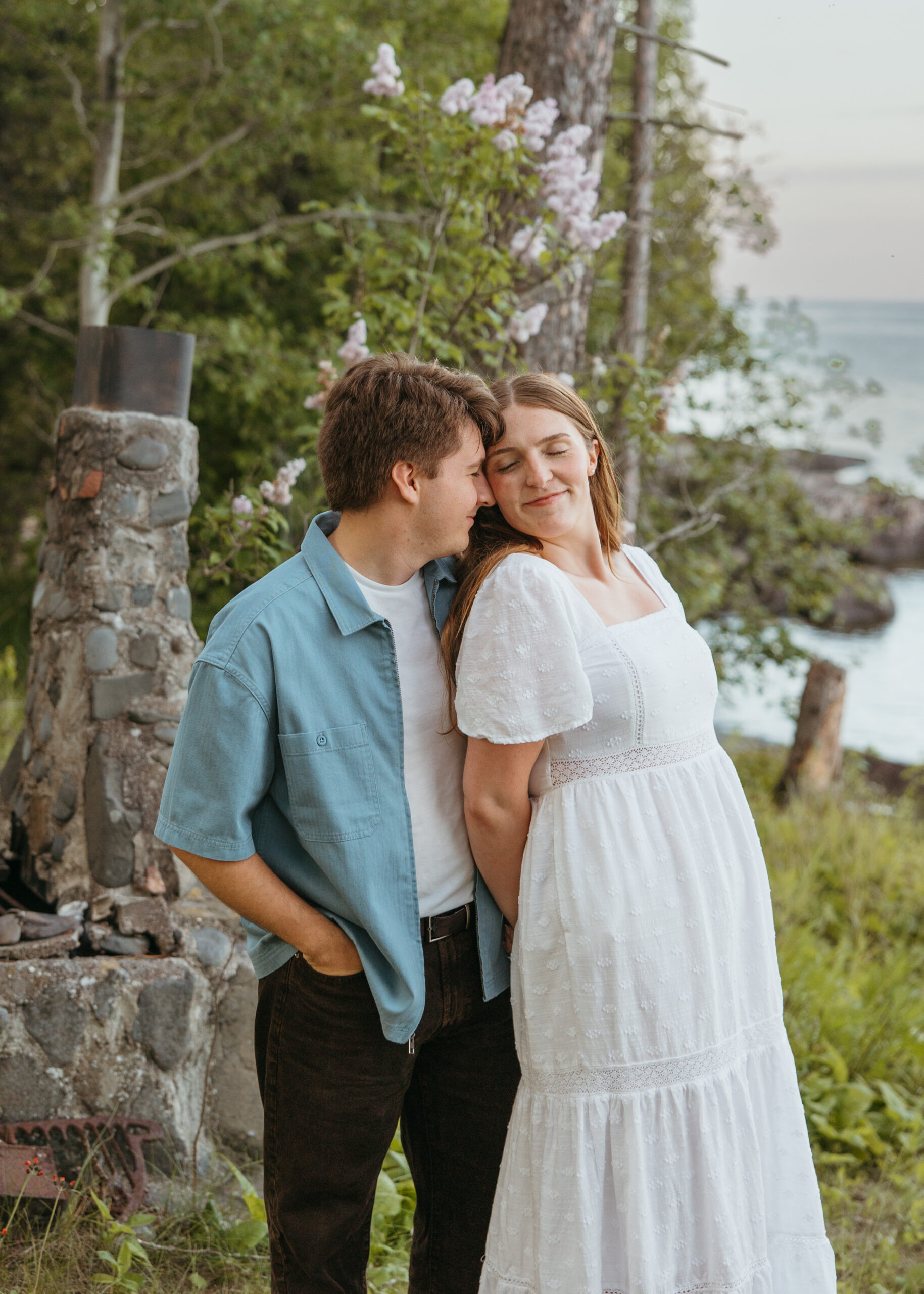 A happy couple standing on the rugged, rocky shoreline of Lake Superior in Lutsen, Minnesota, with waves crashing against the rocks under a bright summer sky.