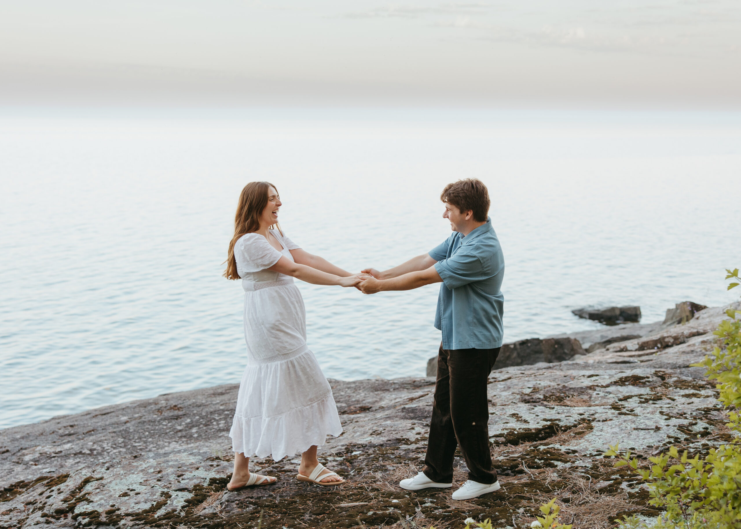 Wide shot of a couple embracing on a natural rock shelf along Lake Superior during an engagement session on the North Shore, surrounded by pine trees and water.