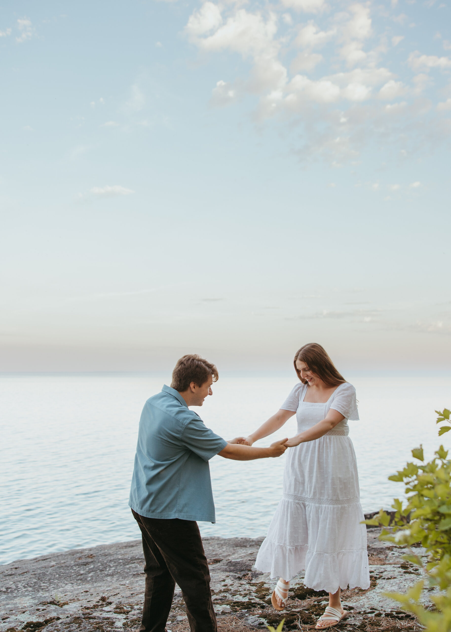 Wide shot of a couple embracing on a natural rock shelf along Lake Superior during an engagement session on the North Shore, surrounded by pine trees and water.