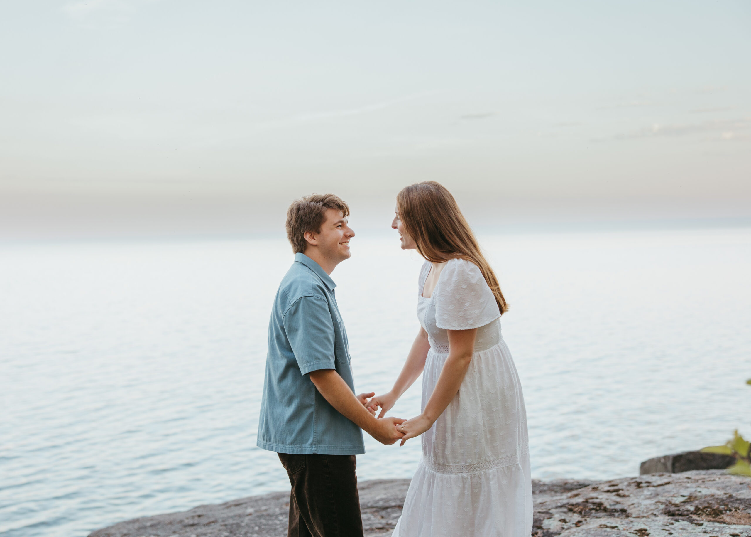 Wide shot of a couple embracing on a natural rock shelf along Lake Superior during an engagement session on the North Shore, surrounded by pine trees and water.
