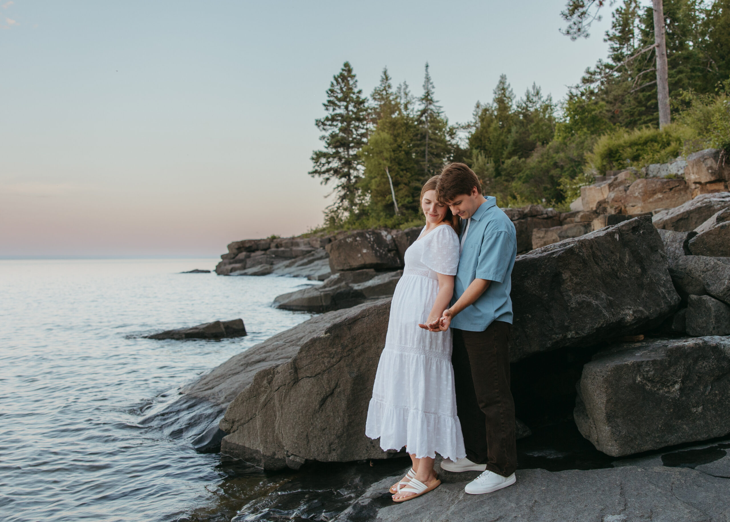 Wide shot of a couple embracing on a natural rock shelf along Lake Superior during an engagement session on the North Shore, surrounded by pine trees and water.