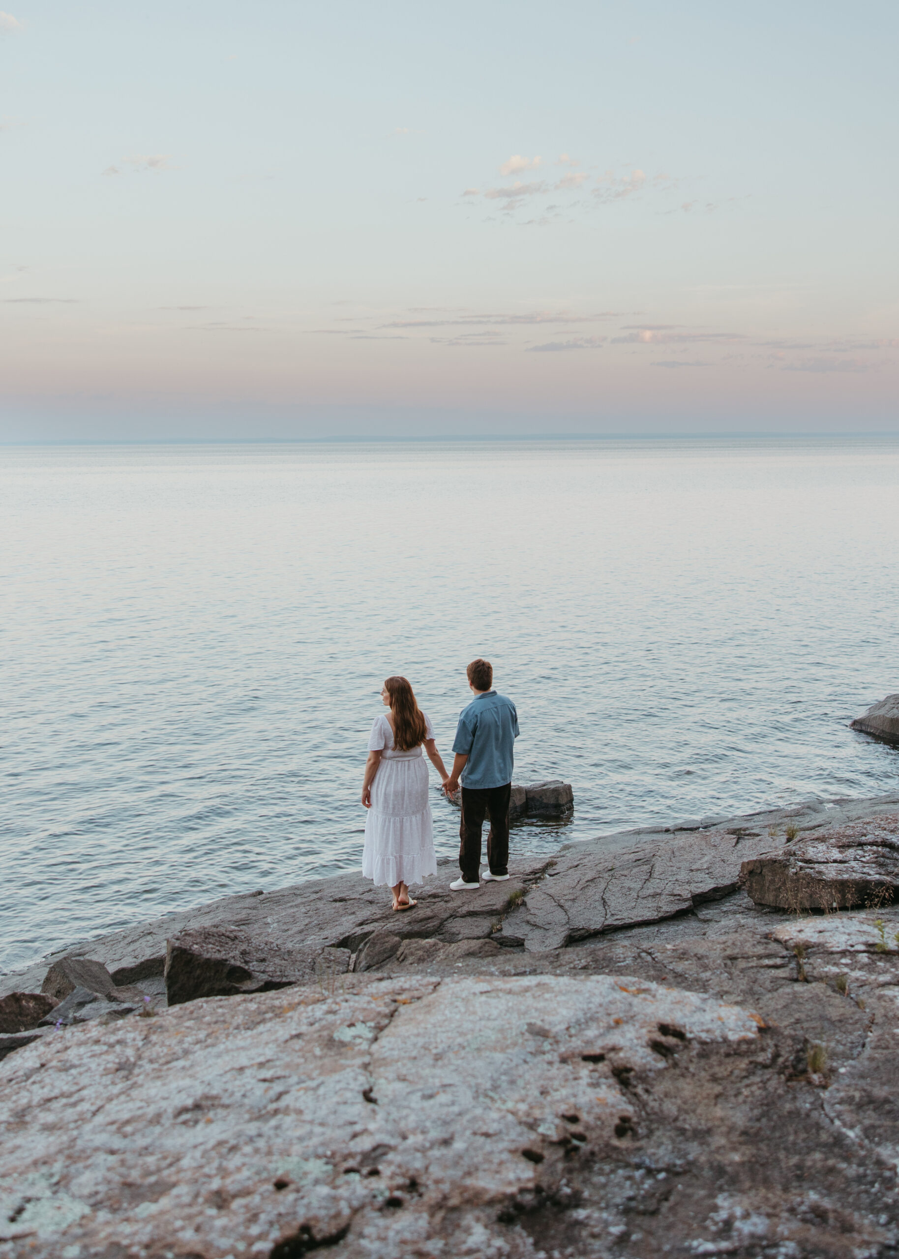 Wide shot of a couple embracing on a natural rock shelf along Lake Superior during an engagement session on the North Shore, surrounded by pine trees and water.